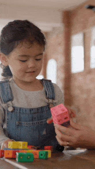A child in denim overalls interacts with colorful cube toys held by an adult.