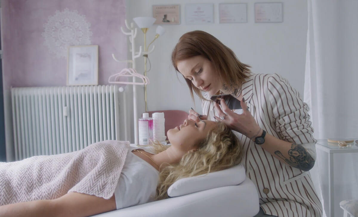 A woman lies on a treatment bed while another woman applies eyelash extensions in a bright, tidy beauty salon.