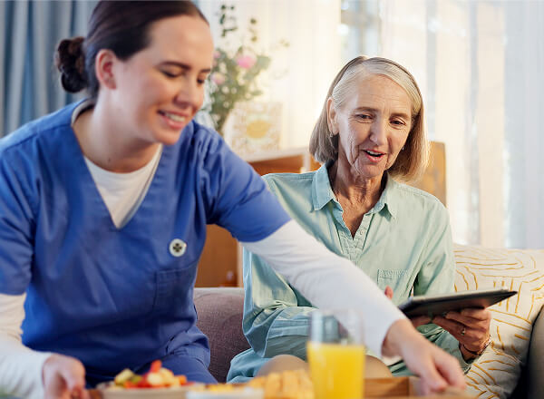 A nurse in blue scrubs serves breakfast to an older woman with gray hair, who is sitting on a couch holding a tablet.