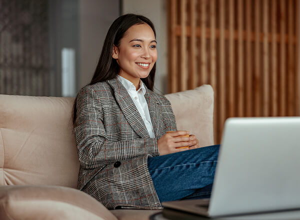 A woman in a plaid blazer sits on a beige sofa, holding a cup and smiling while looking at a laptop in front of her.