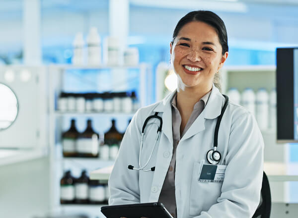 A woman in a white lab coat with a stethoscope around her neck is smiling and holding a tablet in a medical or laboratory setting.