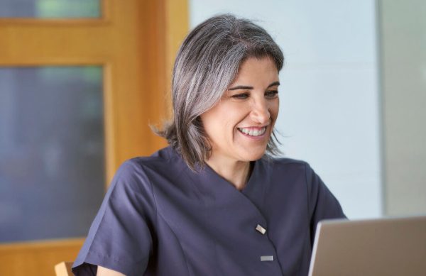 A woman with gray hair wearing a dark gray uniform is smiling while looking at a laptop screen indoors.