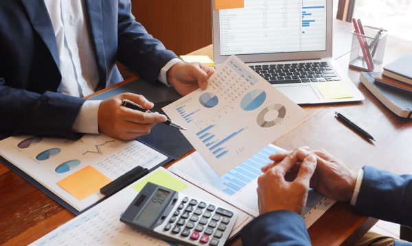 Two people in business attire review financial charts and graphs at a desk with documents, a calculator, and a laptop displaying data.