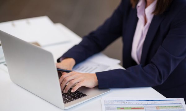 Person in a business suit typing on a laptop at a desk with documents and a spreadsheet visible nearby.
