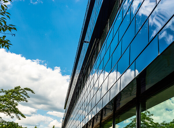 Glass facade of a modern building reflecting blue sky and white clouds, with green trees visible along the edge and in the reflection.