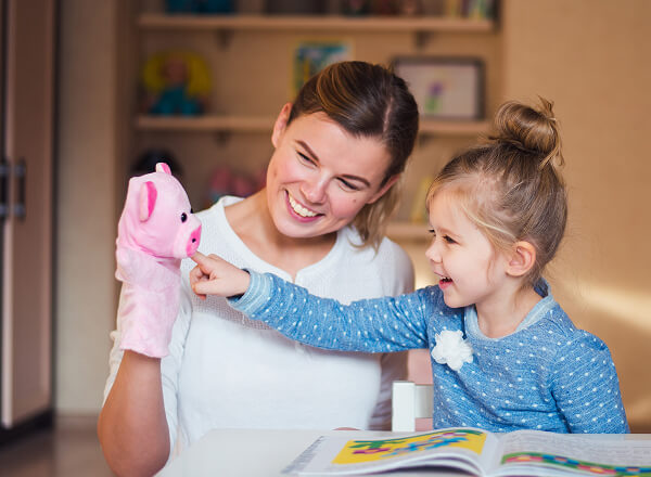 An adult and a young girl sit at a table with an open book. The adult holds a pig hand puppet, and the girl touches the puppet, both smiling.