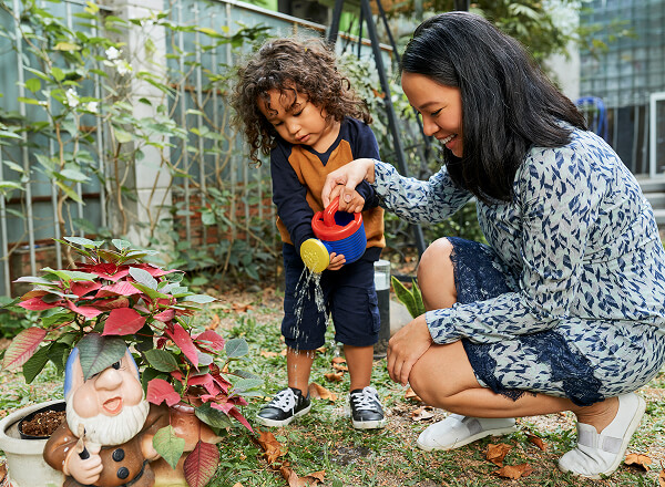 A woman and a young child water a potted plant next to a garden gnome in an outdoor garden area.