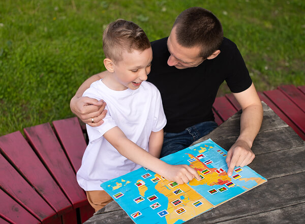 An adult and a child sit at an outdoor table, smiling and pointing at a colorful map or game board with small flag markers.