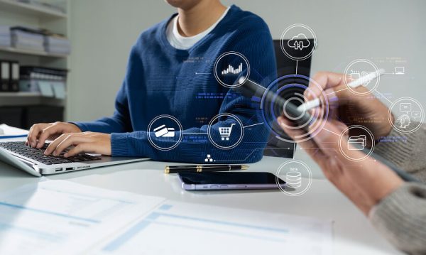 Two people work at a desk with a laptop and papers, while digital finance and technology icons are overlaid in the foreground.