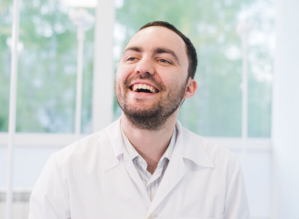 A man with short dark hair and a beard, wearing a white lab coat, smiles and looks slightly upward in a bright room with large windows.