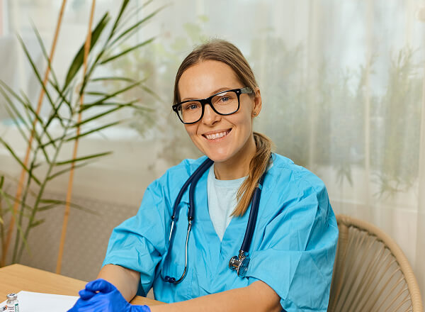 A healthcare professional wearing scrubs, a stethoscope, glasses, and gloves sits at a desk, smiling at the camera.