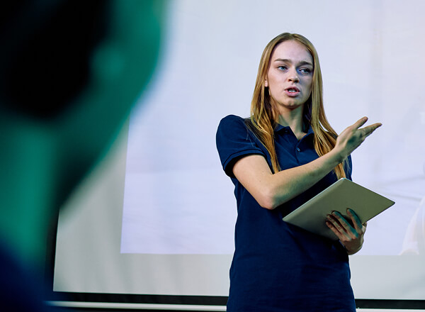 A woman in a navy shirt holds a tablet and gestures while speaking in front of a screen, addressing a blurred audience member.