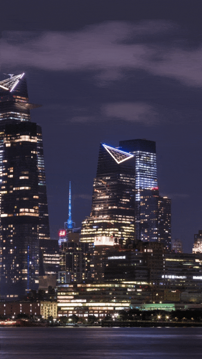 Nighttime cityscape featuring illuminated modern skyscrapers with geometric designs and a glowing tower against a dark sky. River in the foreground reflects lights from the buildings.