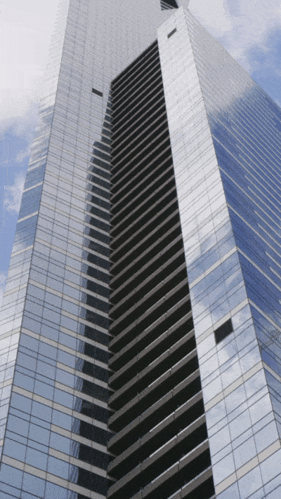 A tall, modern skyscraper with a glass facade reflecting the sky and nearby structures. The building has a central section with black horizontal lines.