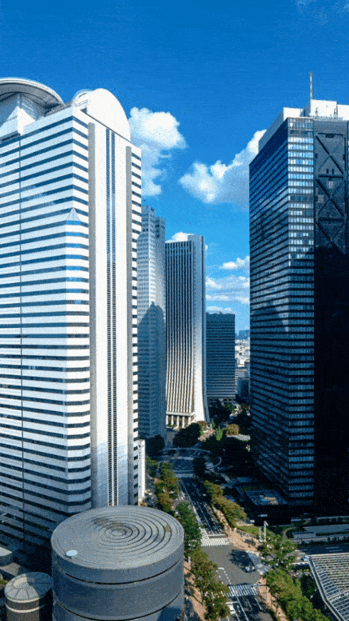 Tall modern skyscrapers against a blue sky with scattered clouds, lining a city street with visible traffic and greenery.