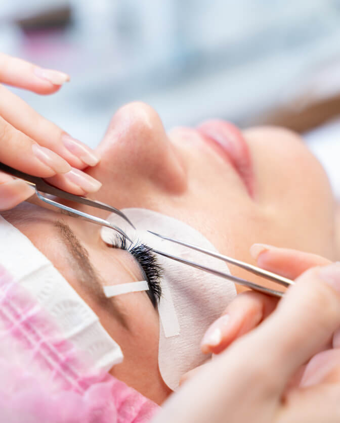 A close-up of a person receiving eyelash extensions, with tweezers applying lashes while eye pads protect the skin.