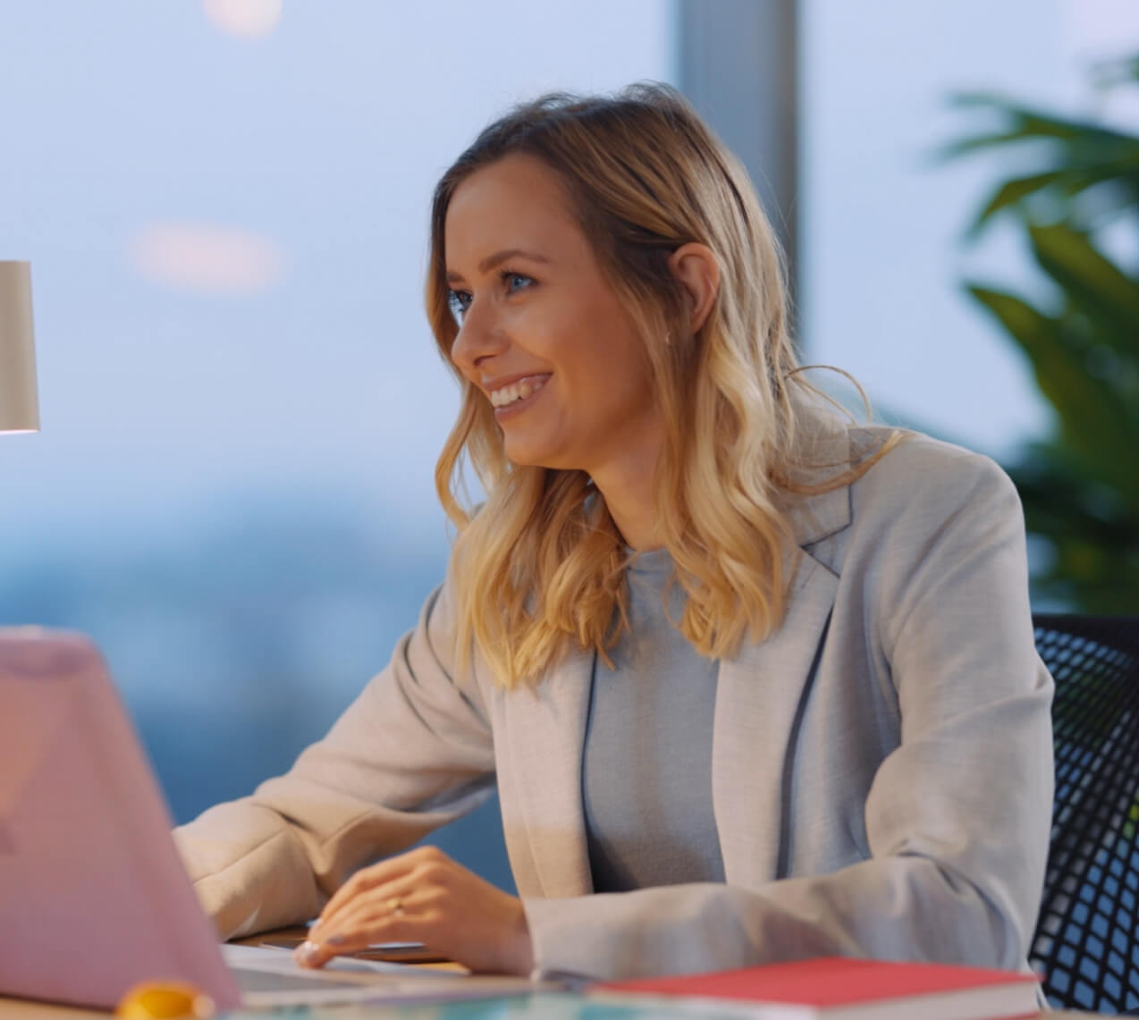 Woman with blonde hair in a light blazer sits at a desk, smiling while using a laptop. There are books and plants in the background.