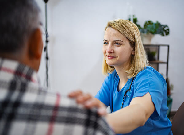 A female healthcare professional in blue scrubs places a comforting hand on a patient's shoulder during a consultation.