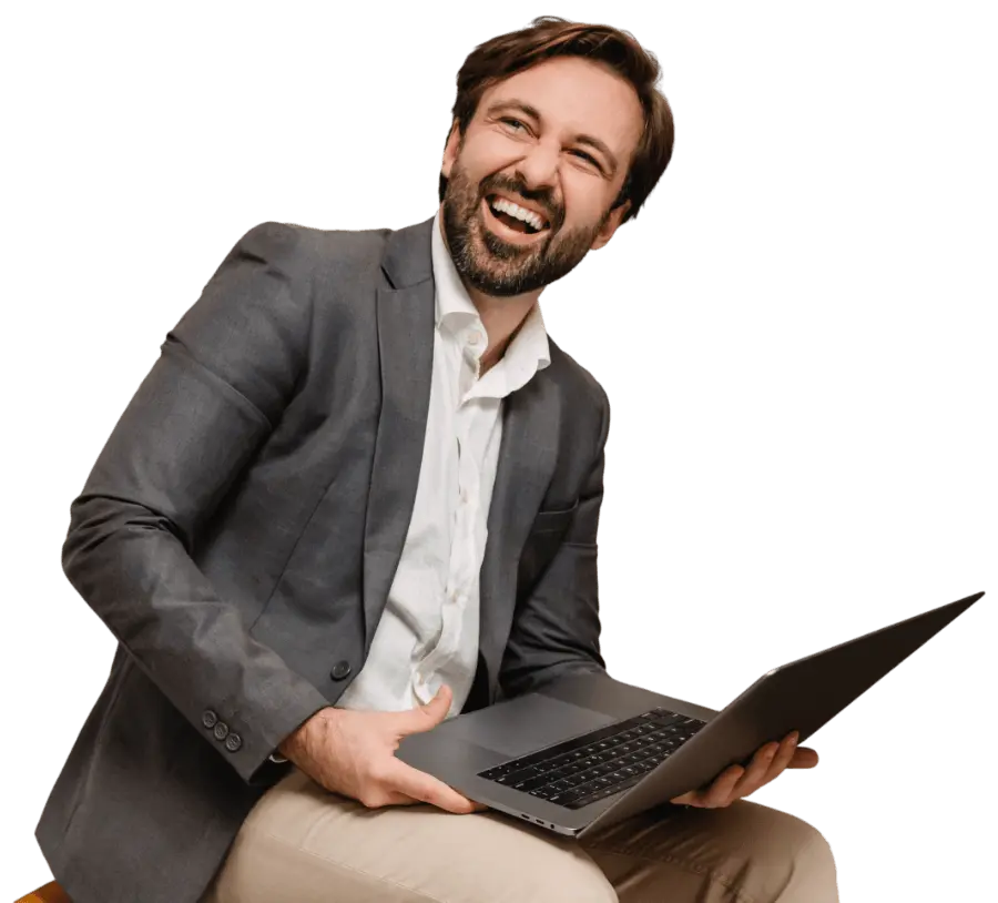 Man in a gray blazer and white shirt sits and smiles broadly while holding an open laptop.