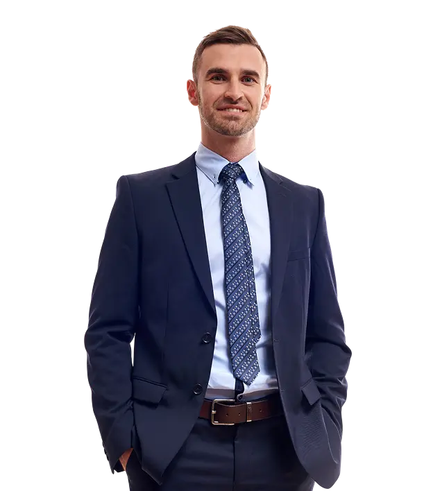 A man wearing a navy blue suit, light blue dress shirt, and patterned tie stands with his hands in his pockets, facing forward and smiling slightly.