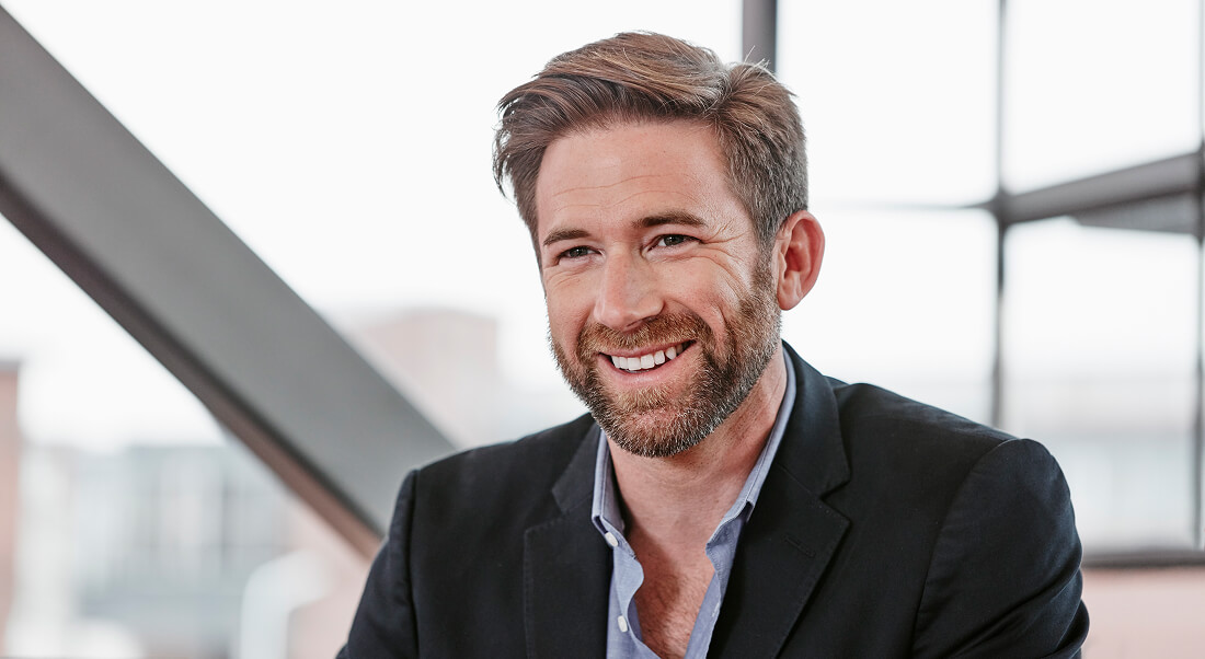 A man with brown hair and a beard, wearing a dark blazer and blue shirt, sits indoors and smiles. Large windows and structural beams are visible in the background.