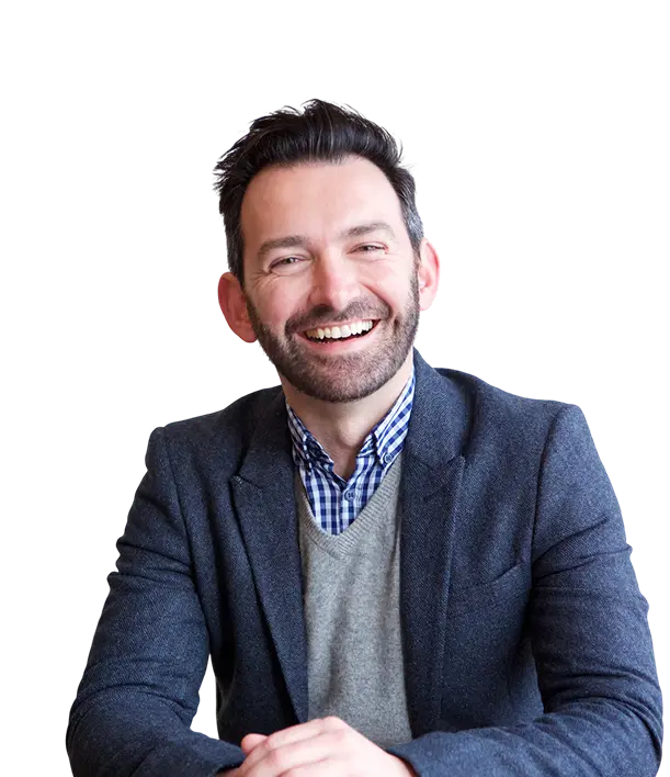 A man with dark hair and a beard, wearing a blazer and checked shirt, sits and smiles at the camera.