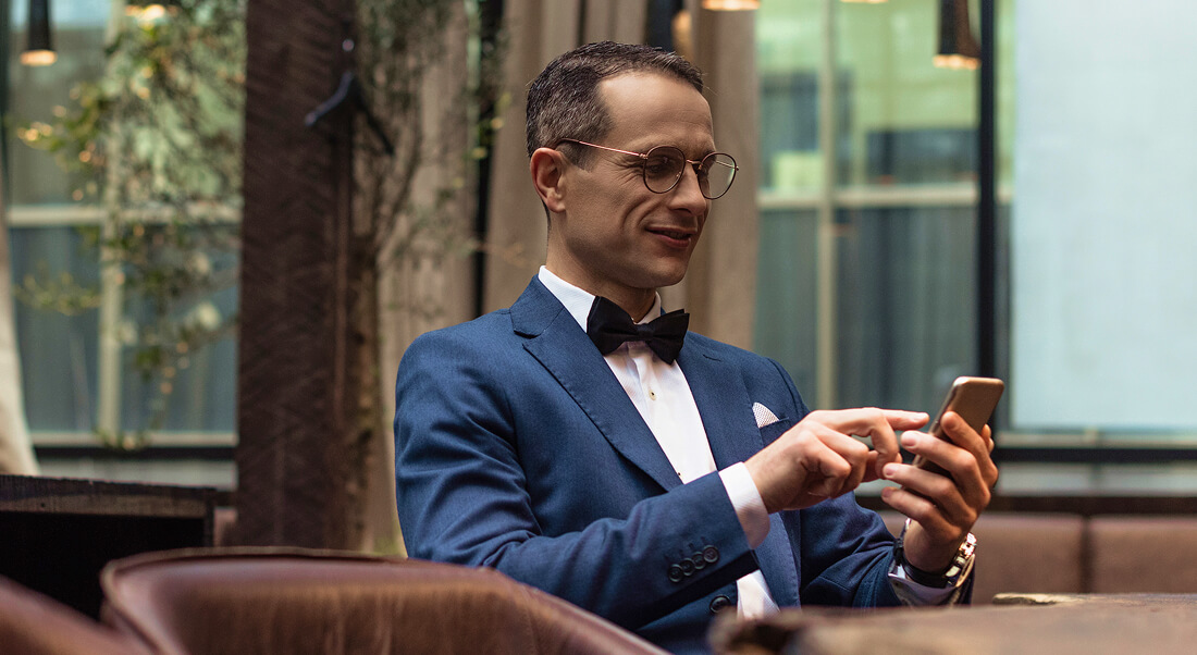 A man in a blue suit and bow tie sits indoors, looking at a smartphone and smiling slightly.