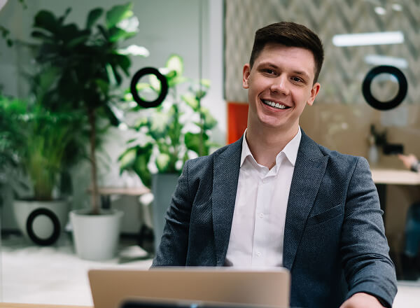 A young man in a suit sits at a desk with a laptop, smiling in a modern office with plants and glass walls in the background.