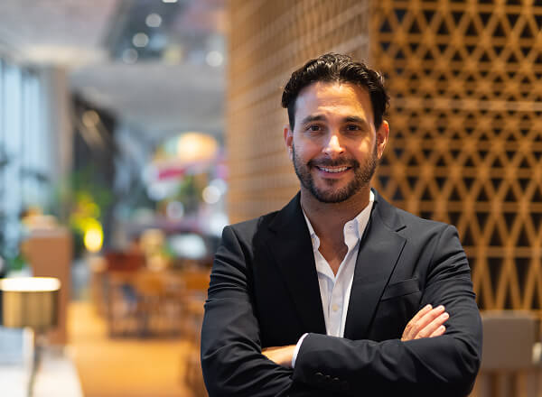 Man in a dark suit stands indoors with arms crossed, smiling at the camera; modern office space with warm lighting in the background.
