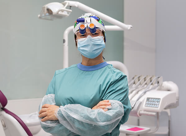 A dentist wearing magnifying loupes, a mask, and a surgical gown stands with arms crossed in a dental clinic.