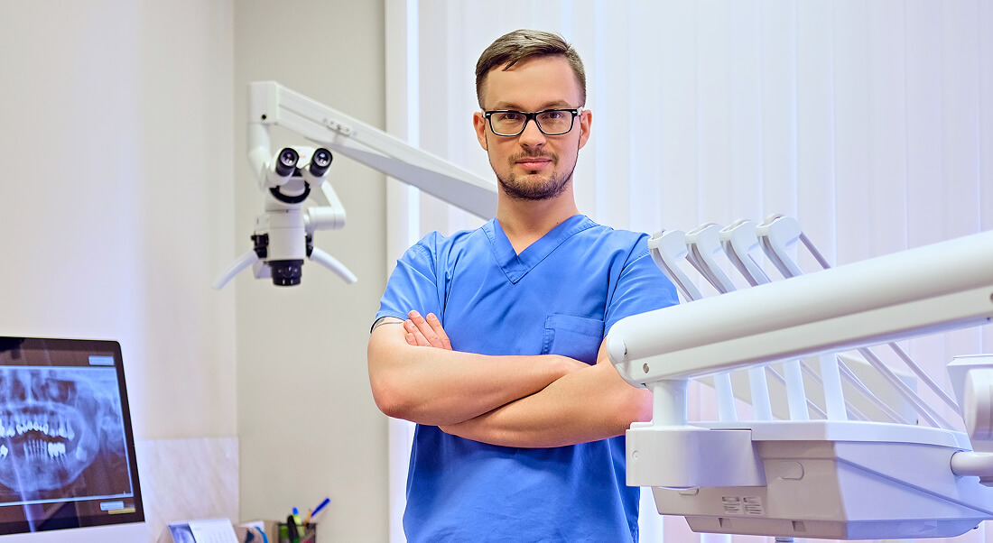 A male dentist in blue scrubs stands with arms crossed in a dental office, surrounded by dental equipment and an X-ray image on a monitor.