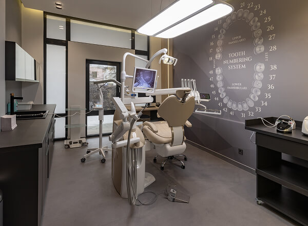 Modern dental clinic room with a dental chair, equipment, monitors, and a tooth numbering system chart on the wall.