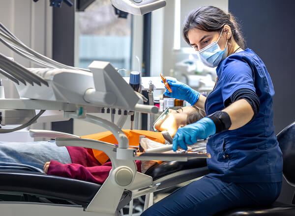 A dentist wearing gloves and a mask prepares dental instruments while a patient lies in the dentist chair, ready for treatment.