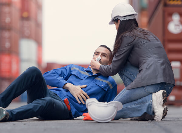 A worker lies on the ground holding his stomach while another person in a hard hat attends to him, possibly providing first aid at an industrial site.
