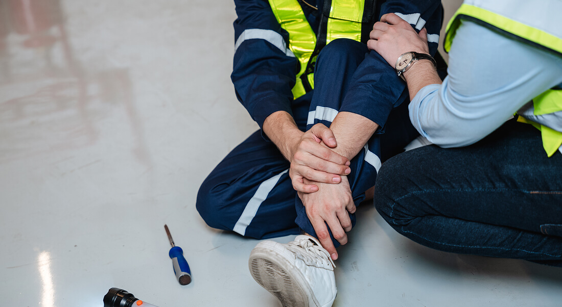 A worker in a reflective vest sits on the floor holding their ankle, while another person offers assistance; tools lie nearby.