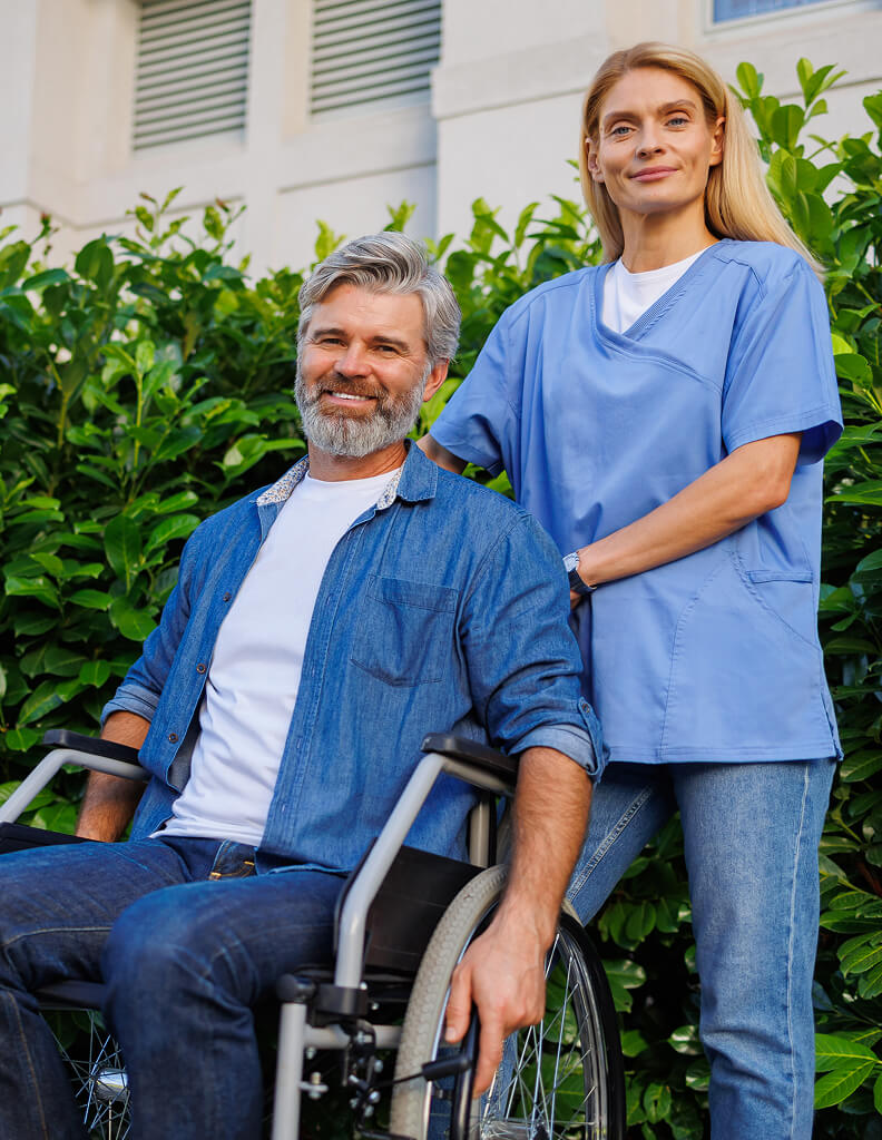 A man in a wheelchair sits outdoors next to a woman in light blue scrubs standing behind him, both looking at the camera and smiling.