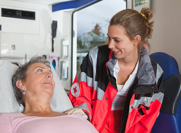 A paramedic in a red jacket attends to a woman lying on a stretcher inside an ambulance.