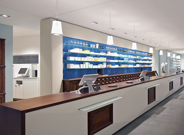 A modern pharmacy interior with a long counter, computer registers, shelves stocked with medication, and overhead lighting.
