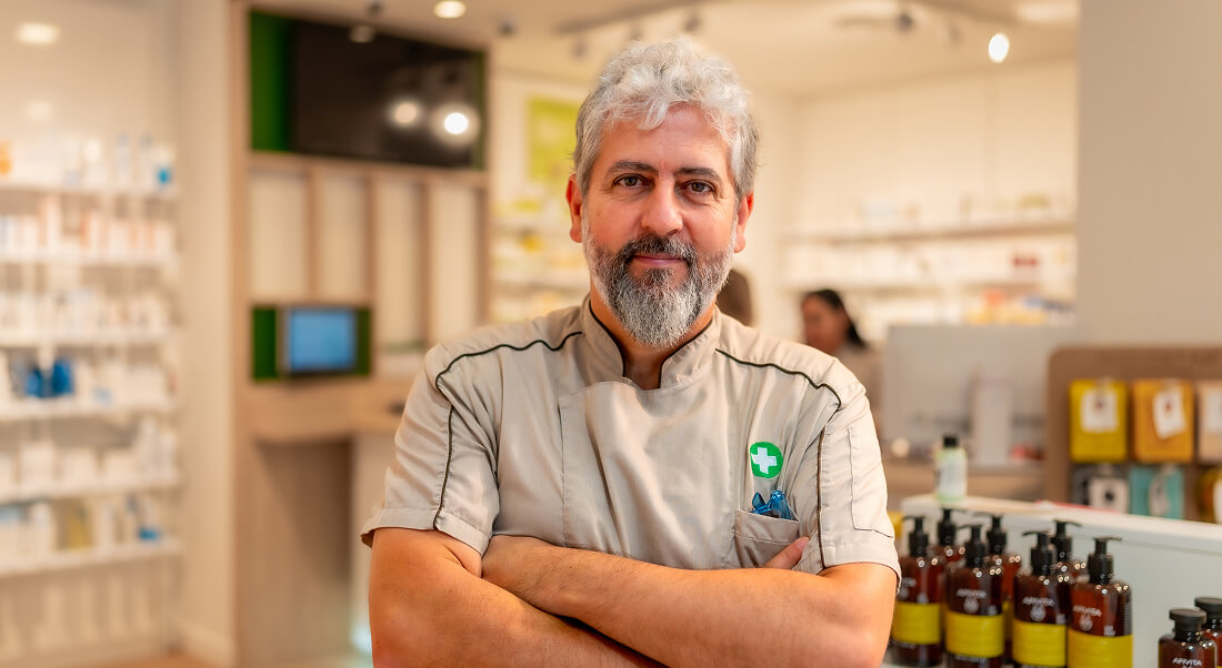 A pharmacist with gray hair and beard stands with arms crossed in front of shelves filled with pharmacy products.