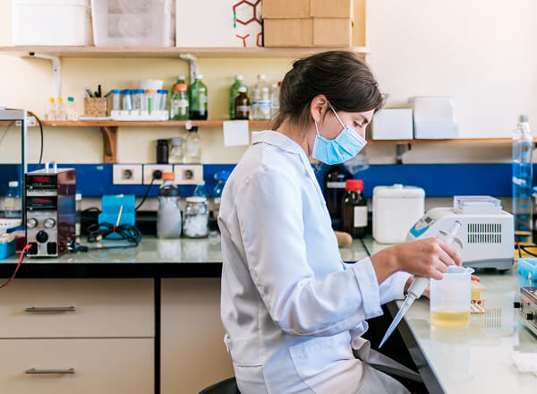 A person wearing a lab coat and face mask uses a pipette at a laboratory workstation with equipment and bottles in the background.