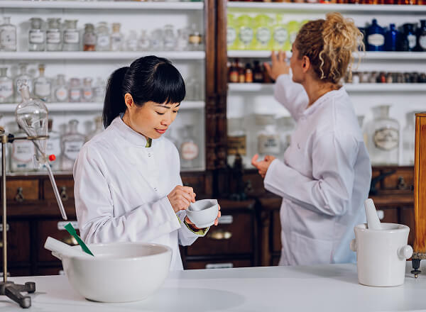 Two pharmacists in white coats work in a pharmacy; one grinds substances in a mortar and pestle while the other reaches for bottles on a shelf behind.