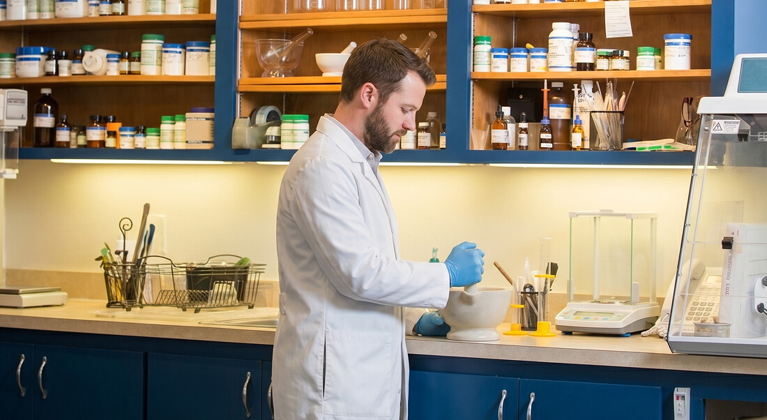 A man in a lab coat and gloves uses a mortar and pestle at a laboratory counter, with shelves of bottles and scientific equipment in the background.