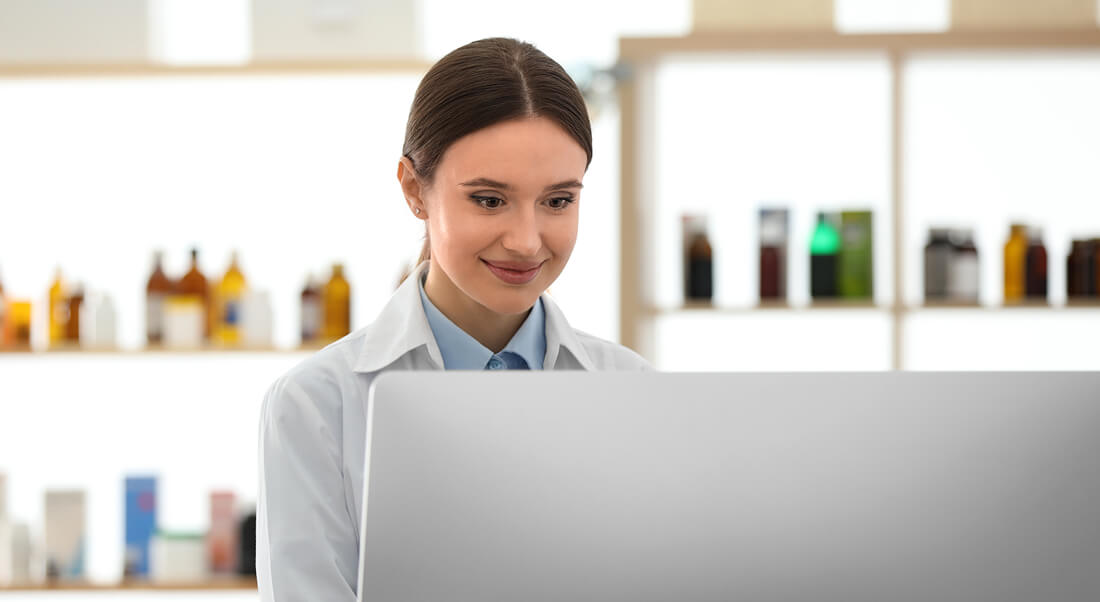 A woman in a lab coat sits at a desk and looks at a computer monitor, with shelves of bottles and containers in the background.