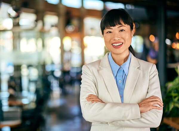 A woman in a light-colored suit stands indoors with arms crossed, smiling confidently. The background is blurred, suggesting a professional or business setting.