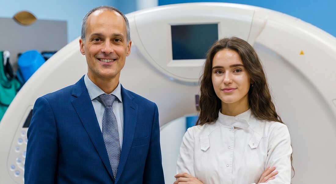 A man in a suit and a woman in a lab coat stand in front of a medical imaging machine in a clinical setting.