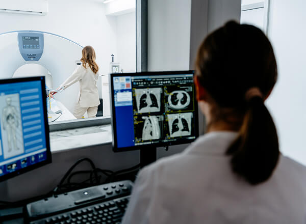 A technician monitors medical scans on computer screens while a patient prepares for a CT scan in an adjacent room.