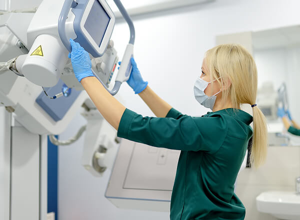 A woman wearing a mask and gloves adjusts a medical imaging machine in a clinical setting.
