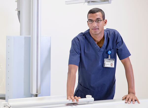 A medical professional in blue scrubs and glasses stands beside medical equipment, wearing an ID badge.