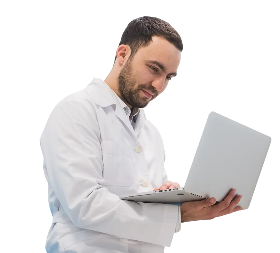 A man in a white lab coat is holding and using a laptop, looking at the screen.