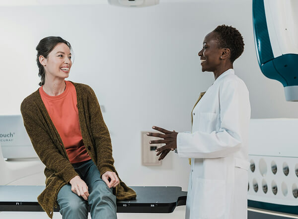 A woman sits on a medical examination table, smiling and talking with a healthcare professional in a white coat in a clinical setting.
